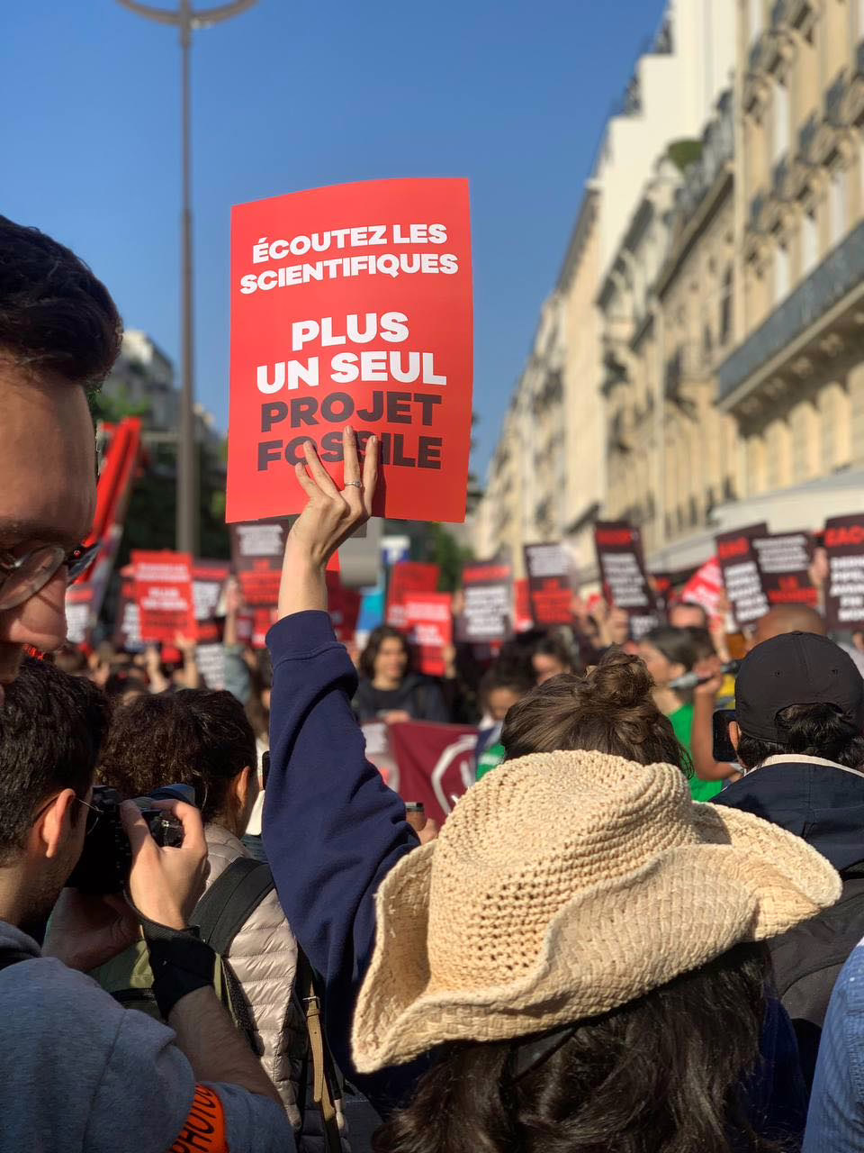 un.e manifestant.e dans un cortège, tenant une pancarte rouge qui se détache sur le ciel bleu, marqué : "écoutez les scientifiques : PLUS UN SEUL PROJET FOSSILE"