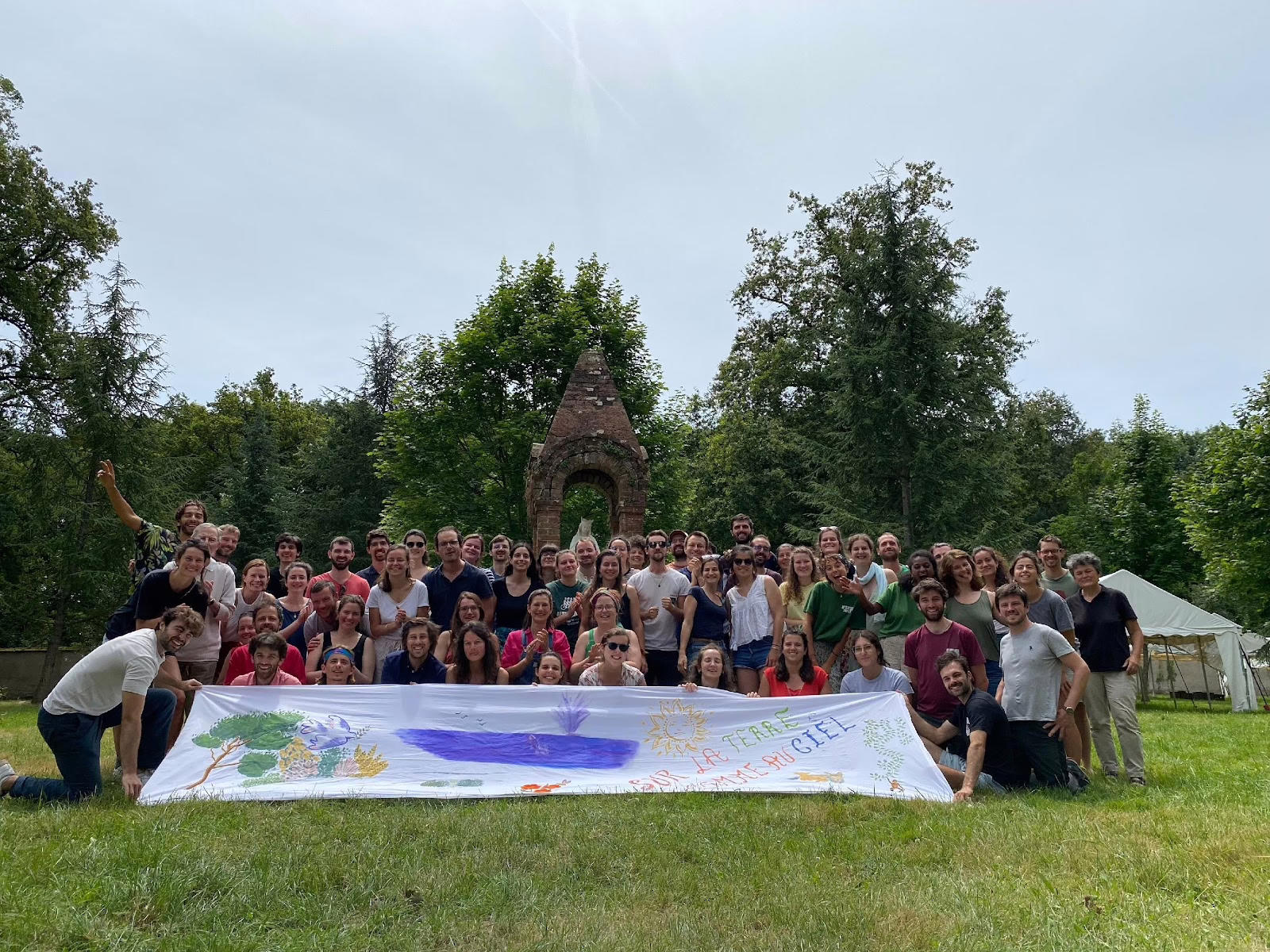 Photo de groupe des participants aux Festiv'été, le festival de Lutte et Contemplation, avec une banderole aussi chouette que peu lisible, on distingue simplement la phrase "sur la terre comme au ciel"